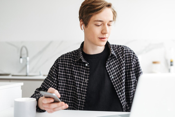 Männliche junge weiße Person mit dunkelblonden mittellangen Haaren sitzt am Küchentisch und blickt auf einen Laptop, zudem hält sie ein Handy in der Hand. Die Person trägt ein schwarzes T-Shirt und kariertes schwarzes Hand was offen getragen wird. 